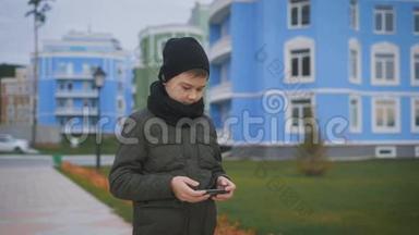 穿着温暖的<strong>深灰</strong>色夹克和黑色帽子和围巾的孩子站在街上玩电话。 男孩正在给